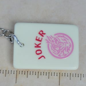 May include: A rectangular, off-white keychain charm with the word "JOKER" in red and a pink graphic. The charm is approximately 2 inches long and has a silver chain attached.