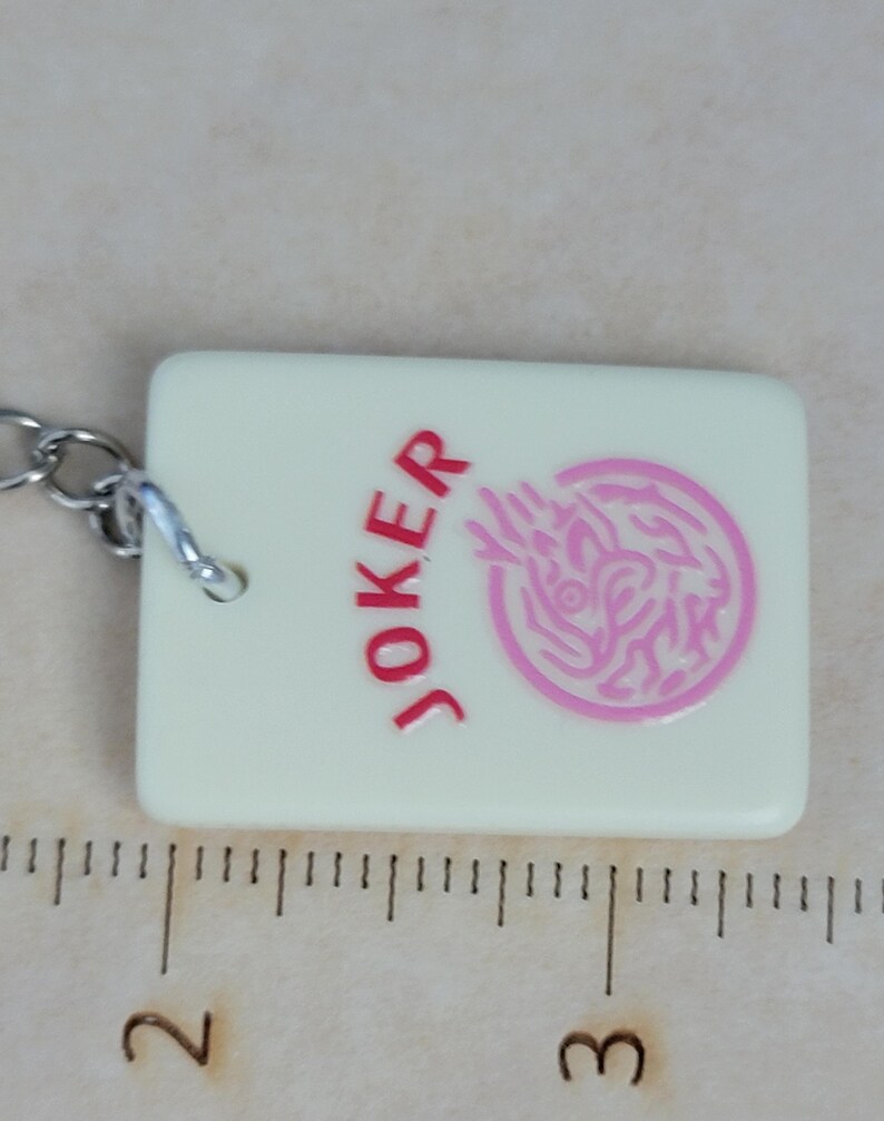 May include: A rectangular, off-white keychain charm with the word "JOKER" in red and a pink graphic. The charm is approximately 2 inches long and has a silver chain attached.