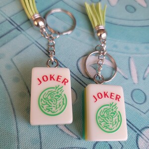 May include: Two keychains with rectangular white charms featuring the word "JOKER" in red and a green dragon design. Each keychain has a silver ring, chain, and a light green tassel.