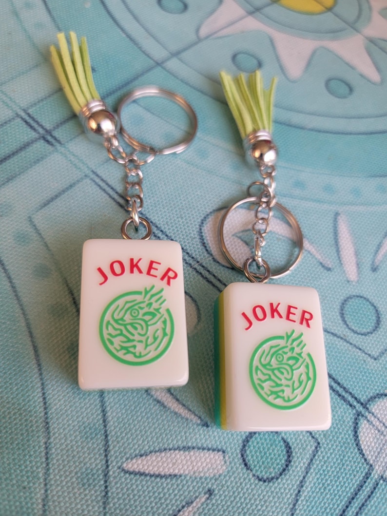 May include: Two keychains with rectangular white charms featuring the word "JOKER" in red and a green dragon design. Each keychain has a silver ring, chain, and a light green tassel.