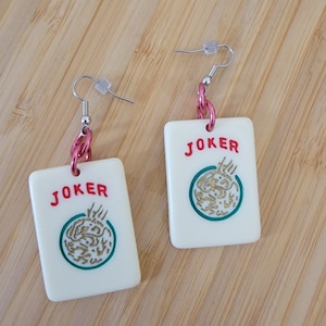 May include: A pair of white, rectangular earrings with a red "JOKER" inscription and a green and gold design. The earrings are attached to pink chains.
