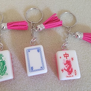 May include: Three keychains with white rectangular charms featuring Chinese characters and colorful illustrations. Each keychain has a silver ring, chain, and a bright pink tassel. The charms have red, green, and blue designs.