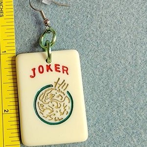 May include: A white ceramic earring with a green and gold design. The earring features the word "JOKER" in red lettering and a green circle with a gold design inside. The earring is attached to a silver hook.