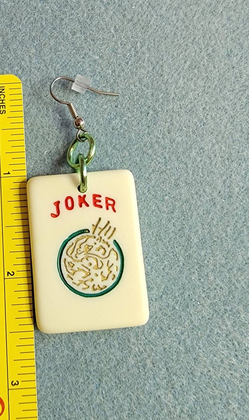 May include: A white ceramic earring with a green and gold design. The earring features the word "JOKER" in red lettering and a green circle with a gold design inside. The earring is attached to a silver hook.