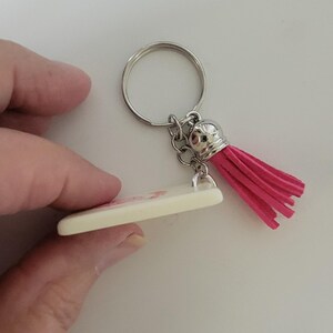May include: A keychain with a silver ring and chain, a cream-colored rectangular charm, and a bright pink tassel. The charm appears to have a pink design. The keychain is held by a person.
