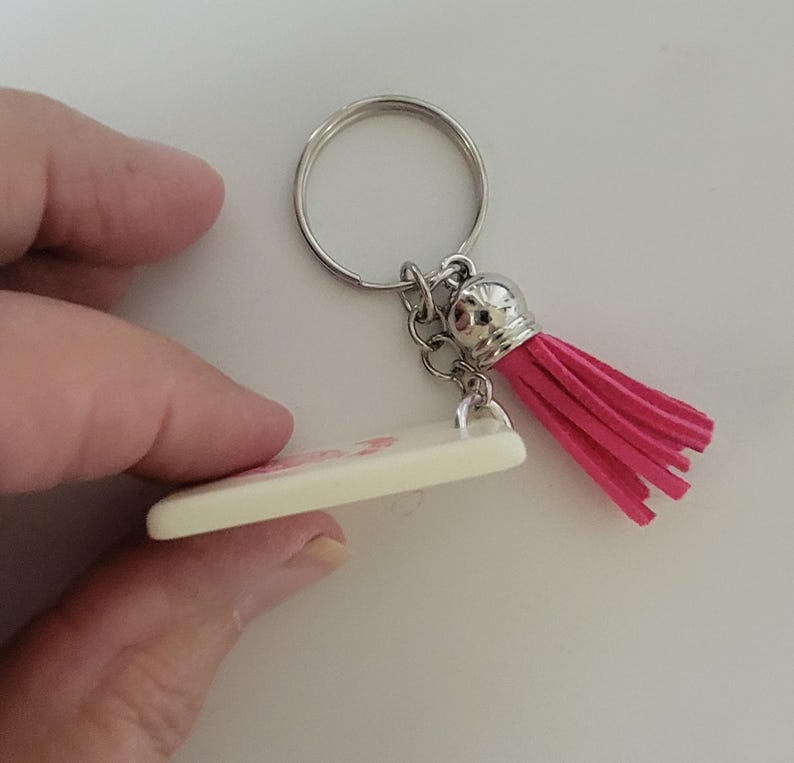 May include: A keychain with a silver ring and chain, a cream-colored rectangular charm, and a bright pink tassel. The charm appears to have a pink design. The keychain is held by a person.