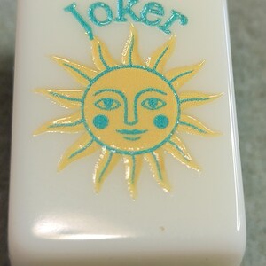 May include: An off-white rectangular Mahjong tile featuring the word "Joker" in teal script above a yellow sun with a smiling face and teal accents. The bottom edge has yellow and teal stripes.