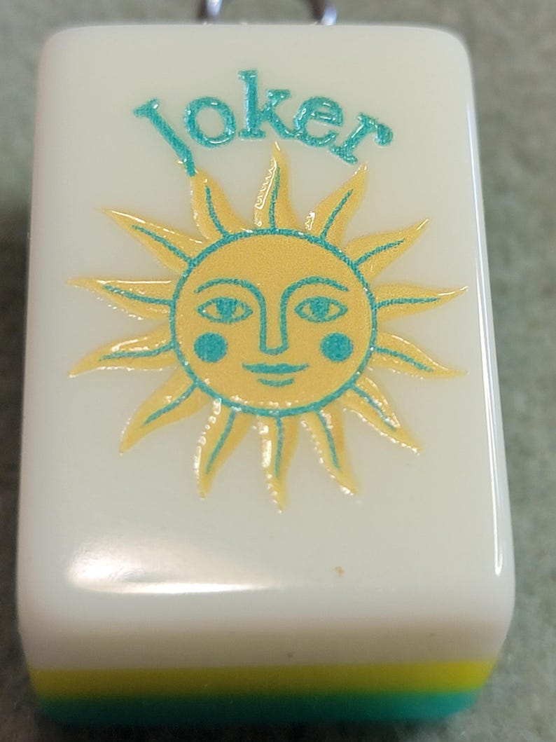 May include: An off-white rectangular Mahjong tile featuring the word "Joker" in teal script above a yellow sun with a smiling face and teal accents. The bottom edge has yellow and teal stripes.