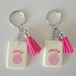 May include: Two keychains with rectangular cream-colored pendants. Each pendant has the word "JOKER" in red and a pink design. The keychains have silver rings and chains, and pink tassels.