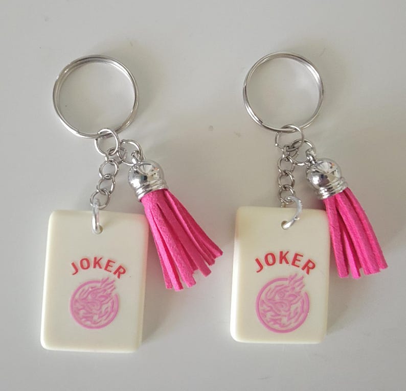 May include: Two keychains with rectangular cream-colored pendants. Each pendant has the word "JOKER" in red and a pink design. The keychains have silver rings and chains, and pink tassels.