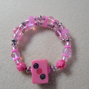 May include: A pink beaded bracelet with a rectangular charm featuring three circular designs. The bracelet has clear and pink beads, silver accents, and pink flower-shaped end pieces. The bracelet is approximately 7 inches in circumference.