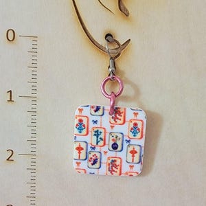 May include: A square pendant earring featuring a white background with a repeating pattern of colorful mahjong tile illustrations in red, blue, and orange. The pendant hangs from a pink metal jump ring and a silver-toned hook, positioned next to a ruler.