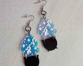 May include: A pair of dangle earrings featuring a black cauldron design with a blue and white cloud-like top. The cloud has white swirls and star-like shapes. The earrings have silver-colored hooks and black connecting rings.