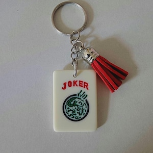May include: A white keychain with a red tassel. The keychain features a white rectangular tile with the word "JOKER" printed in red and a green and black design.