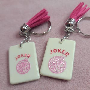 May include: Two keychains with white rectangular pendants featuring the word "JOKER" in pink and a pink graphic. Each keychain has a silver ring, a silver metal accent, and a pink tassel.