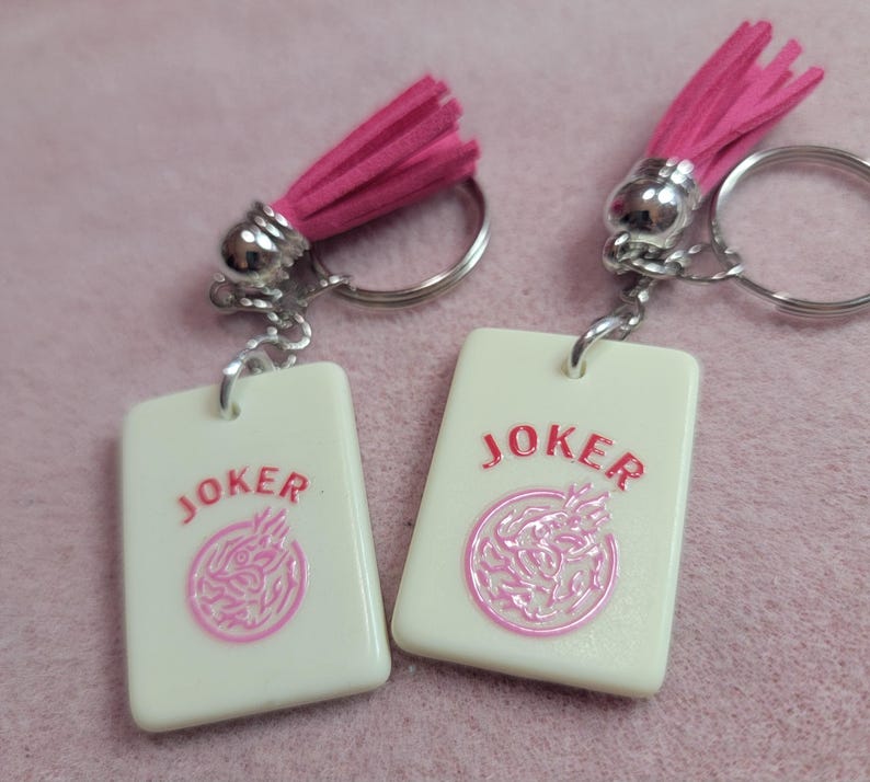 May include: Two keychains with white rectangular pendants featuring the word "JOKER" in pink and a pink graphic. Each keychain has a silver ring, a silver metal accent, and a pink tassel.