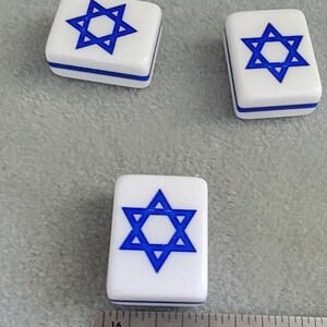 May include: Three white rectangular game tiles with a blue Star of David design. The tiles have a blue border and are arranged on a gray surface. A ruler is in the foreground.