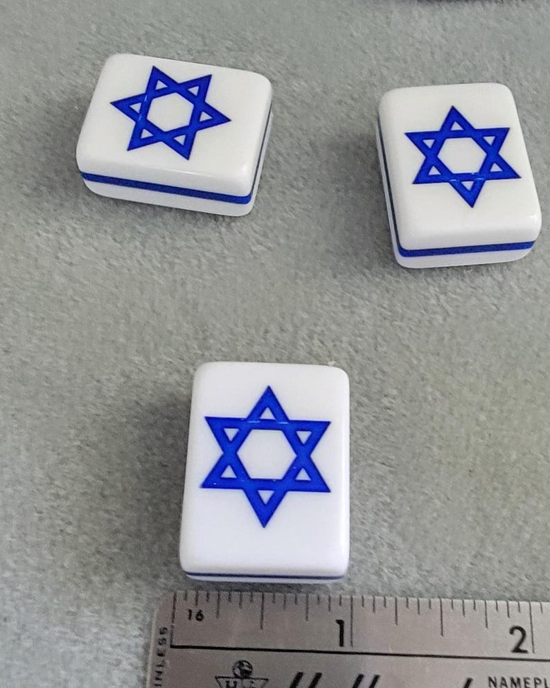May include: Three white rectangular game tiles with a blue Star of David design. The tiles have a blue border and are arranged on a gray surface. A ruler is in the foreground.