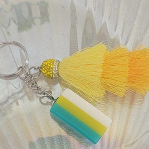 May include: A decorative keychain featuring a yellow and orange tassel, a yellow rhinestone-studded ball, and a rectangular charm with blue, yellow, and white stripes. The keychain has a silver ring and chain.