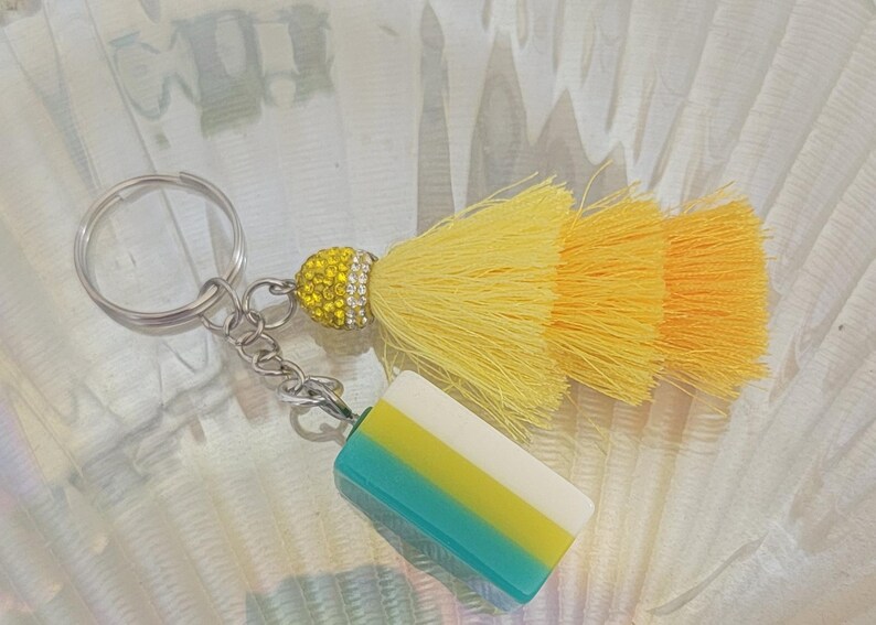 May include: A decorative keychain featuring a yellow and orange tassel, a yellow rhinestone-studded ball, and a rectangular charm with blue, yellow, and white stripes. The keychain has a silver ring and chain.