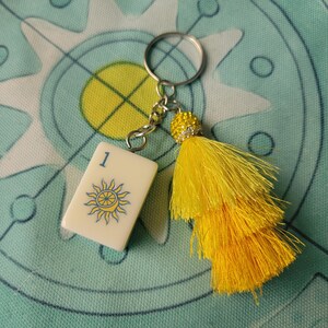 May include: A keychain featuring a white tile with a blue and yellow sun design and the number 1. The keychain has a silver ring, a yellow tassel, and a decorative yellow bead. The background is a teal and white patterned fabric.