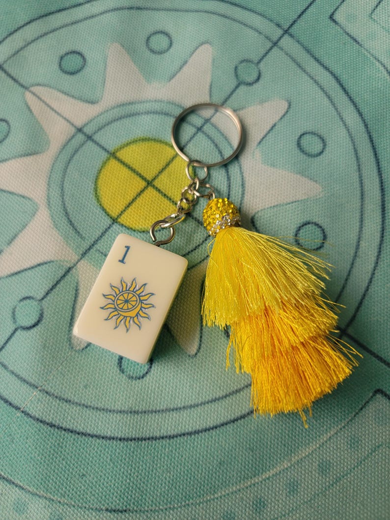May include: A keychain featuring a white tile with a blue and yellow sun design and the number 1. The keychain has a silver ring, a yellow tassel, and a decorative yellow bead. The background is a teal and white patterned fabric.