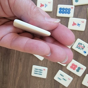 May include: A hand holding a white tile with a blue and green design. The tile is a mahjong tile with the number 9 on it. The tile is surrounded by other mahjong tiles.