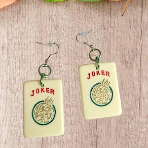 May include: A pair of white earrings with a green and gold design. The earrings are shaped like playing cards with the word "JOKER" printed in red on each card.
