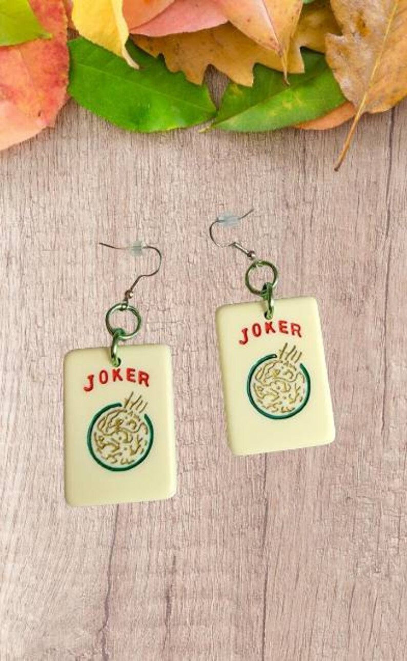 May include: A pair of white earrings with a green and gold design. The earrings are shaped like playing cards with the word "JOKER" printed in red on each card.