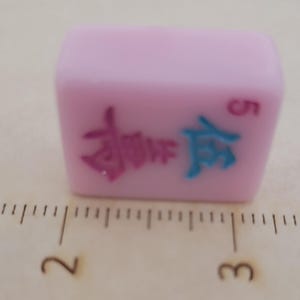 May include: A light pink Mahjong tile with Chinese characters in pink and blue, and the number 5 in red. The tile is rectangular and sits on a surface with measurement markings.