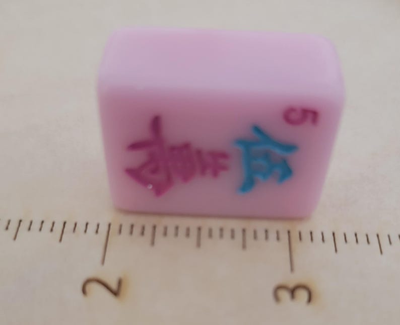 May include: A light pink Mahjong tile with Chinese characters in pink and blue, and the number 5 in red. The tile is rectangular and sits on a surface with measurement markings.