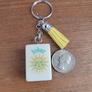 May include: A white keychain charm with the word "Joker" and a sun design in turquoise and yellow. The charm has a yellow tassel and a silver keyring. A coin is next to the charm.