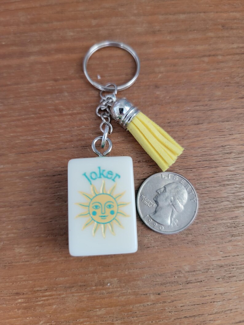May include: A white keychain charm with the word "Joker" and a sun design in turquoise and yellow. The charm has a yellow tassel and a silver keyring. A coin is next to the charm.
