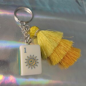 May include: A silver keychain with a white tile featuring a sun design and the number 1. The keychain has a yellow tassel and a decorative yellow beaded accent. The tile has a blue and yellow sun design.