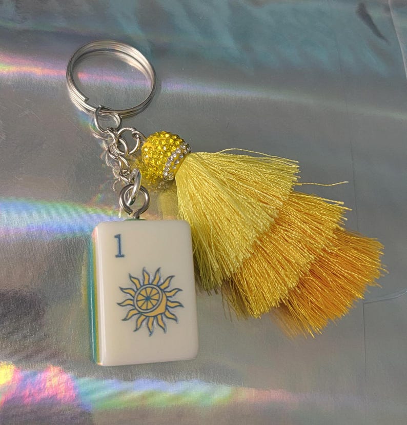 May include: A silver keychain with a white tile featuring a sun design and the number 1. The keychain has a yellow tassel and a decorative yellow beaded accent. The tile has a blue and yellow sun design.