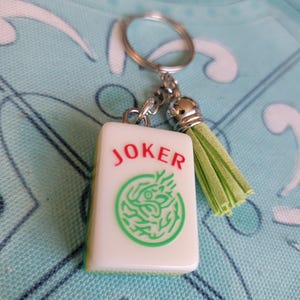 May include: A white Mahjong tile keychain with the word "JOKER" in red and a green dragon design. The keychain has a silver ring, chain, and a light green tassel.