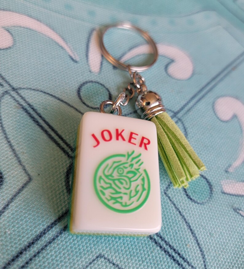 May include: A white Mahjong tile keychain with the word "JOKER" in red and a green dragon design. The keychain has a silver ring, chain, and a light green tassel.