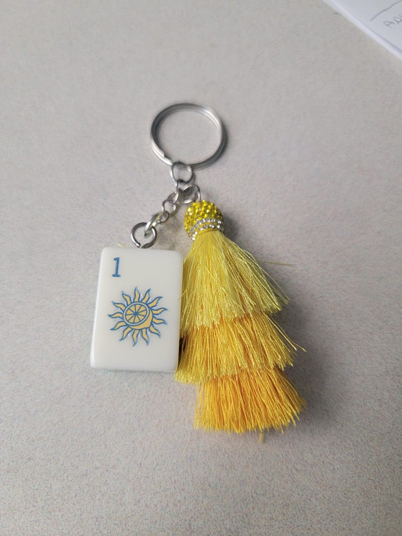 May include: A keychain featuring a white tile with the number 1 and a blue sun design, connected to a yellow tassel and a silver keyring. The tassel is made of layered strands and has a decorative, jeweled top.