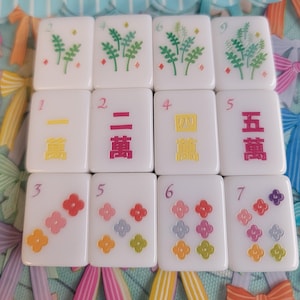 May include: A set of twelve white rectangular mahjong tiles arranged in a grid. The top row features green plant illustrations. The middle row displays Chinese characters in yellow and pink. The bottom row shows colorful flower patterns.