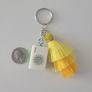 May include: A white keychain charm with a blue and yellow sun design and the number 1. The charm has a yellow tassel and a silver keyring. A quarter is next to the charm.