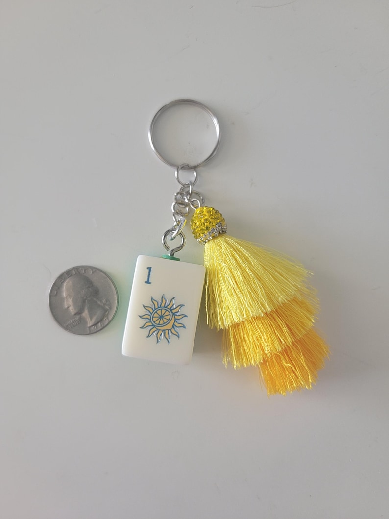 May include: A white keychain charm with a blue and yellow sun design and the number 1. The charm has a yellow tassel and a silver keyring. A quarter is next to the charm.