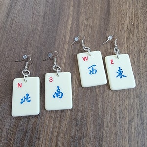 May include: Four pairs of white mahjong tile earrings with red and blue lettering. The tiles are labeled with the directions North, South, East, and West.