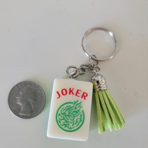 May include: A white rectangular keychain charm with the word "JOKER" in red and a green dragon design. The keychain has a silver ring and a light green tassel. A quarter is next to the keychain.