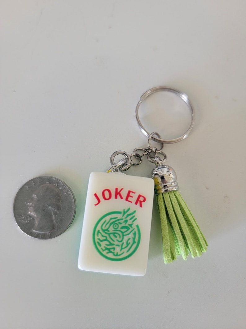 May include: A white rectangular keychain charm with the word "JOKER" in red and a green dragon design. The keychain has a silver ring and a light green tassel. A quarter is next to the keychain.