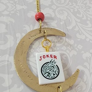 May include: A gold crescent moon wind chime with a red bead, a gold chain, and a small bell. A white card with the word "JOKER" and a black and white design hangs from the crescent moon.