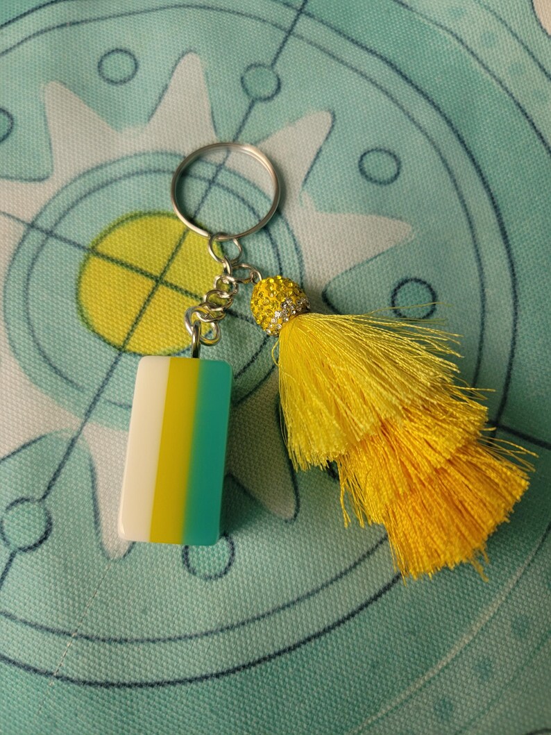 May include: A decorative keychain with a rectangular charm featuring vertical stripes of white, yellow, and teal. It has a silver chain and ring, and a bright yellow tassel with a beaded accent. The background is a teal fabric with a circular design.