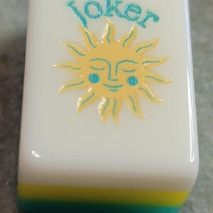 May include: A white Mahjong tile featuring the word "Joker" in teal script above a smiling sun design. The sun is yellow with teal accents. The bottom edge has layers of yellow and teal.