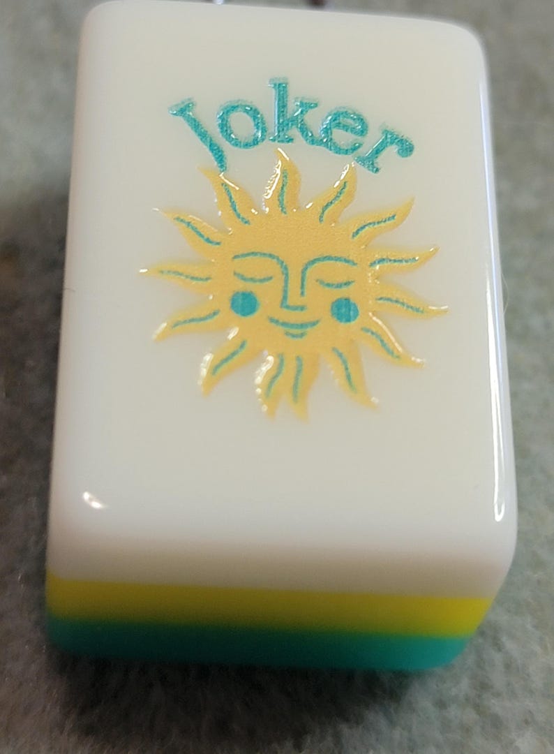 May include: A white Mahjong tile featuring the word "Joker" in teal script above a smiling sun design. The sun is yellow with teal accents. The bottom edge has layers of yellow and teal.