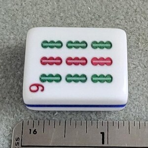 May include: A white Mahjong tile with green and red markings, and the number 6 in red. The tile has a blue stripe along the bottom edge. A metal ruler is in the foreground.