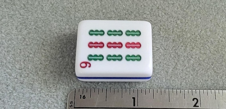 May include: A white Mahjong tile with green and red markings, and the number 6 in red. The tile has a blue stripe along the bottom edge. A metal ruler is in the foreground.